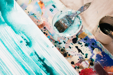 A closeup of used paint brushes sitting on a palette and soaking in water next to a drying piece of abstract art