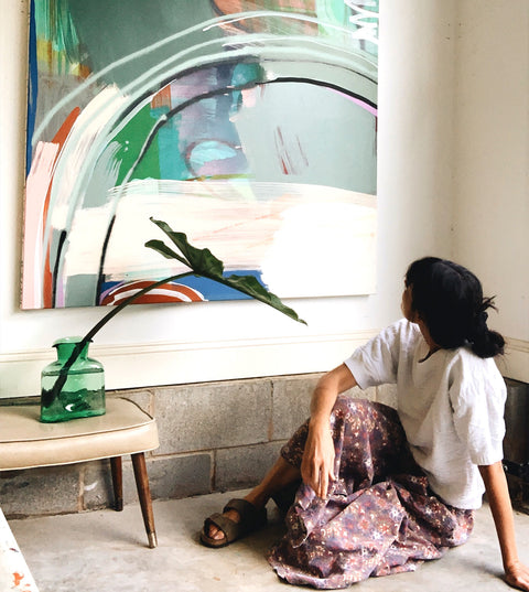 Eva in her studio sitting on the floor looking up at a hanging piece of canvas art mounted on the wall next to her