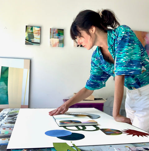 Eva leaning on a desk as she arranges paper cutouts according to a reference in her sketchbook
